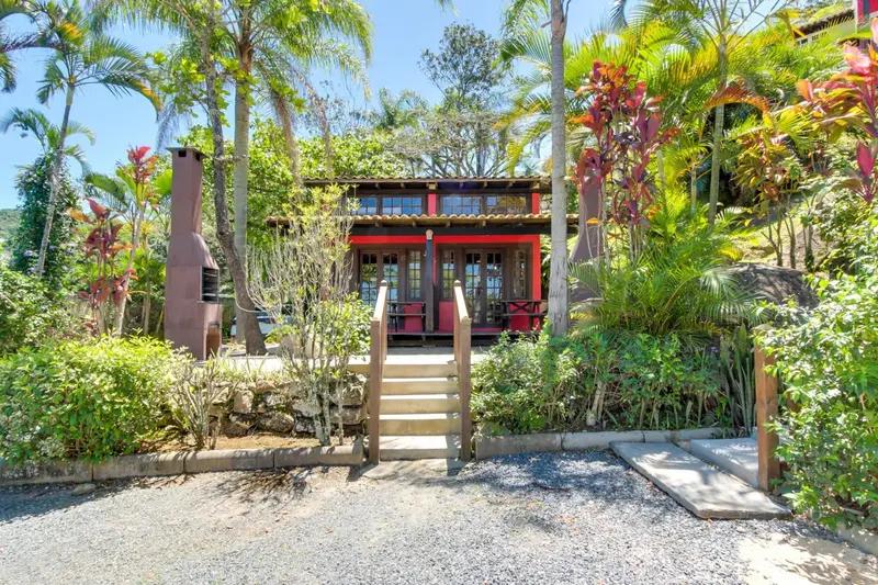 Petite maison rouge avec des escaliers menant à une terrasse en bois, entourée d'une végétation luxuriante.