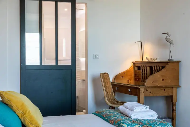 Une chambre lumineuse avec un bureau en bois et une porte vitrée ouverte sur la salle de bain.