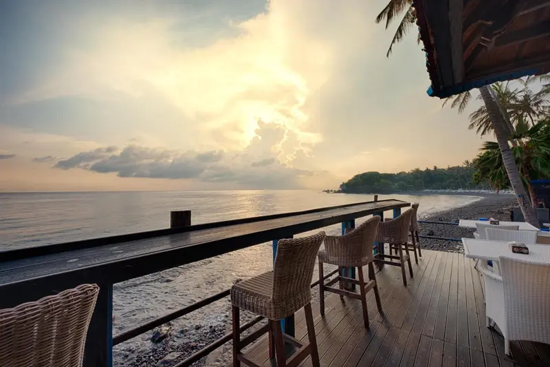 Terrasse en bois avec chaises et tables face à la mer au coucher du soleil.