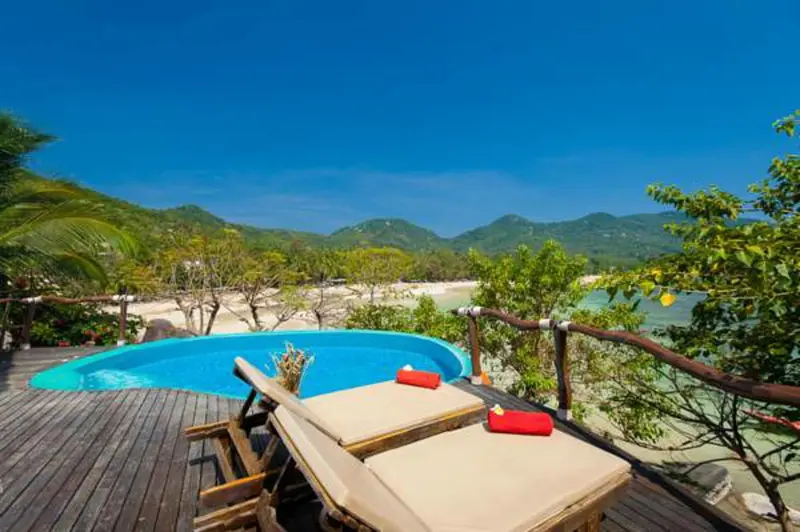 Terrasse en bois avec transats et piscine face à une plage de sable.