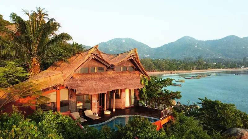 Bungalow en bois avec toit de chaume surplombant une plage et des montagnes.
