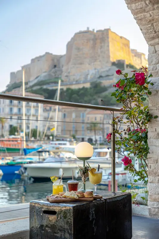 Une table basse avec des boissons et des viennoiseries est mise en scène sur une terrasse avec vue sur un port de plaisance et une forteresse.