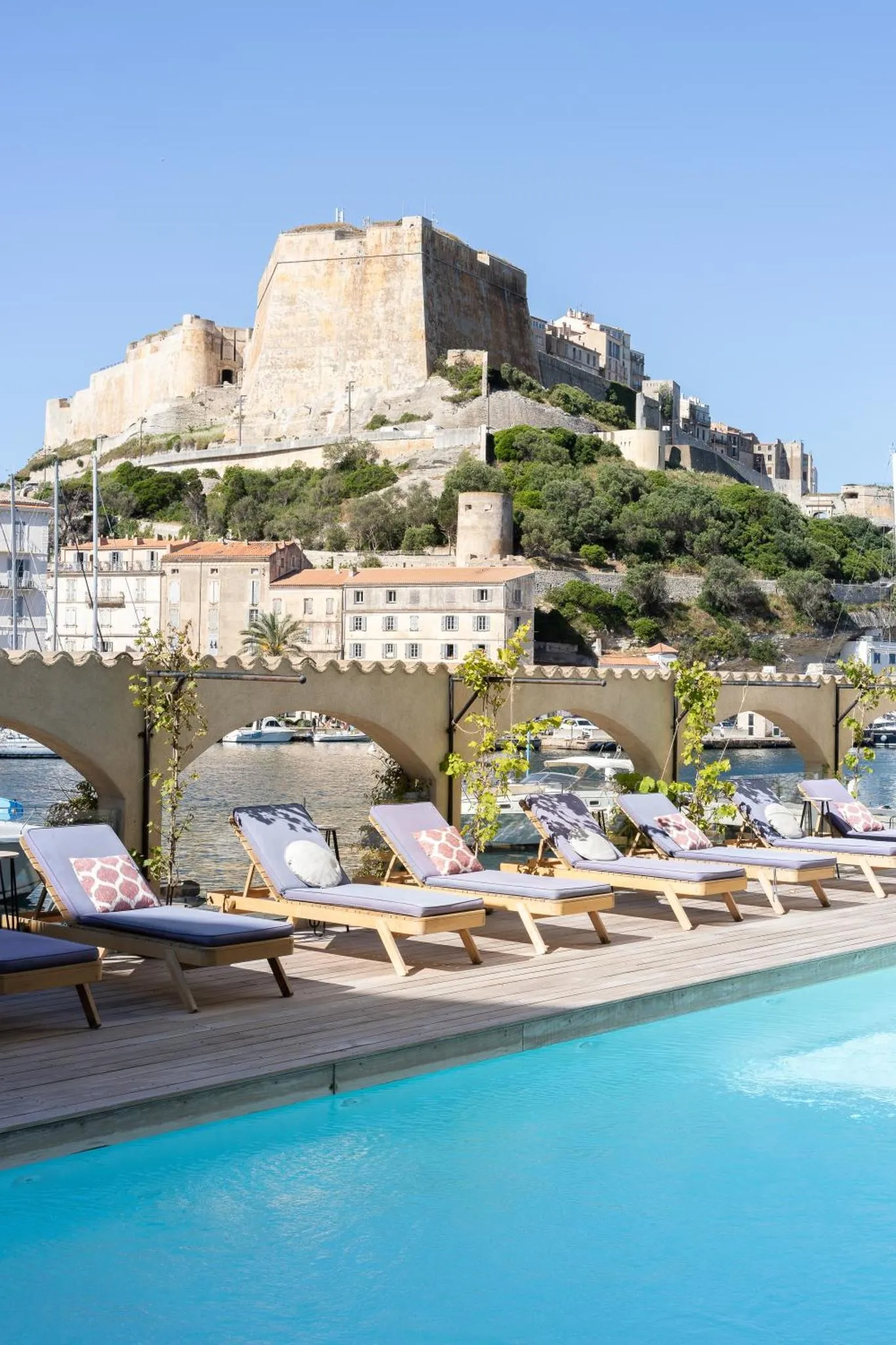 Transats bleus alignés au bord d'une piscine turquoise avec vue sur une ville côtière et un château fort.