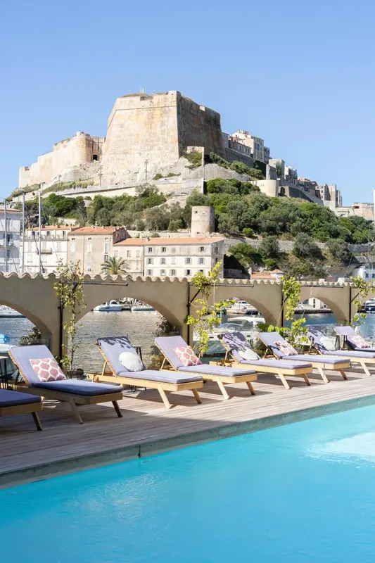 Transats bleus alignés au bord d'une piscine turquoise avec vue sur une ville côtière et un château fort.