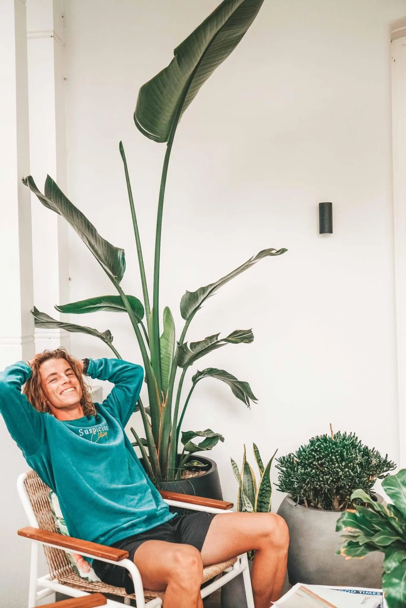 Un homme souriant se détend dans un fauteuil en osier près de plantes tropicales.