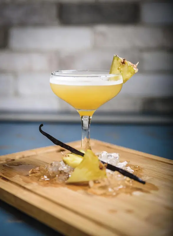 Un cocktail jaune dans un verre à martini, décoré d'un morceau d'ananas et d'une gousse de vanille, est présenté sur une planche en bois avec des glaçons.
