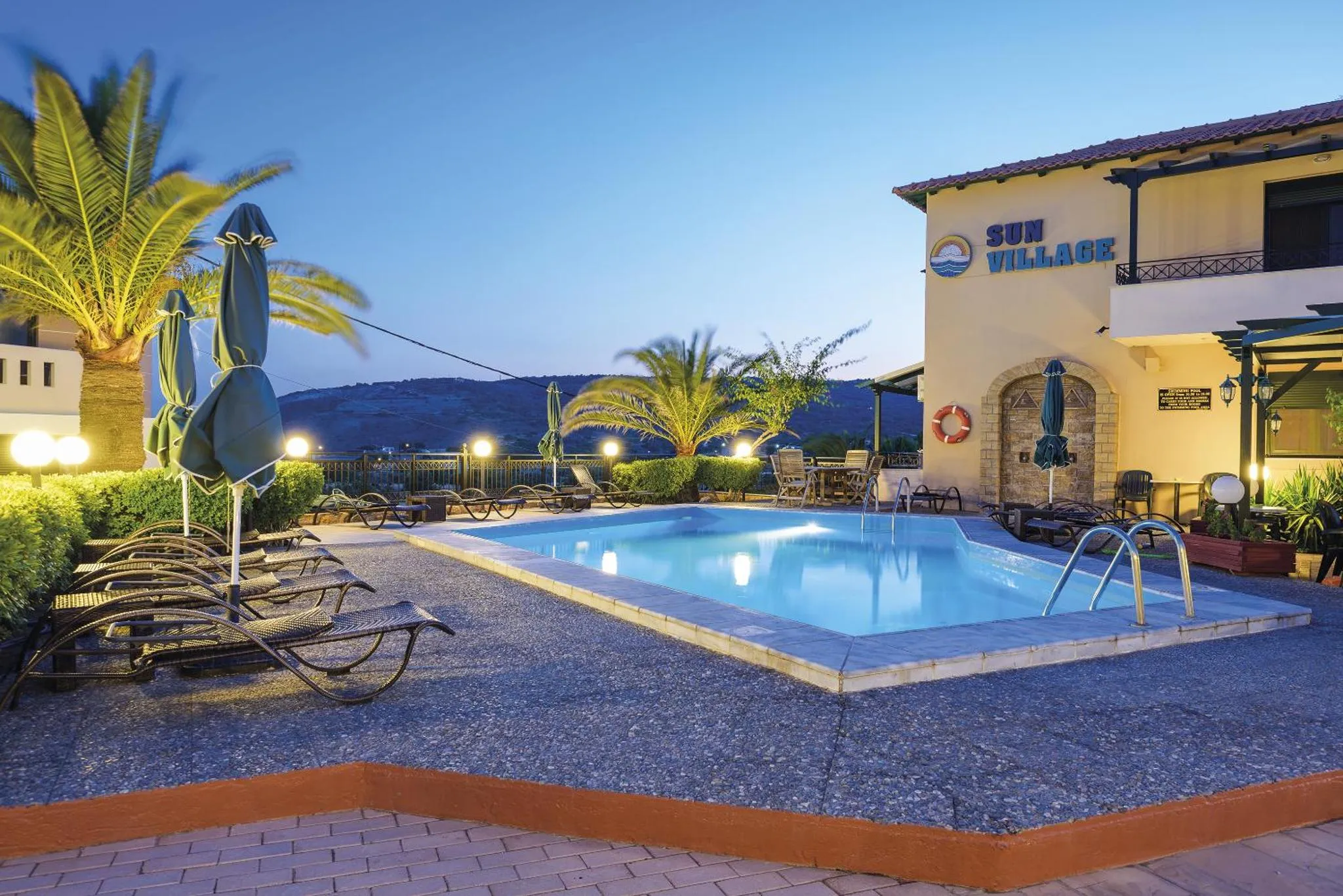 Piscine rectangulaire devant un bâtiment clair avec des transats et des palmiers.