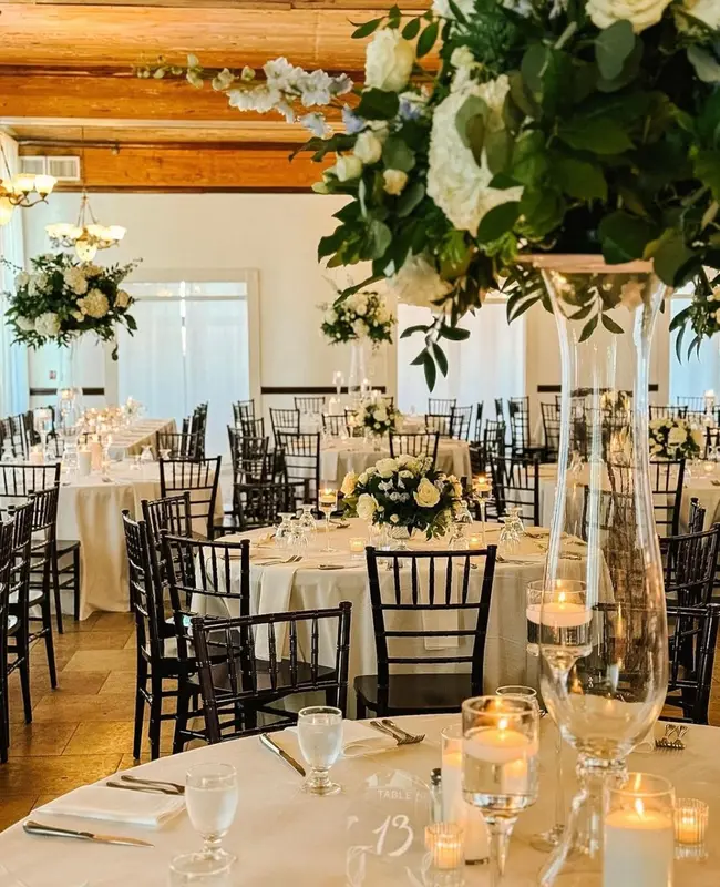 Une salle lumineuse est mise en scène pour un événement avec des tables rondes nappées et des compositions florales.