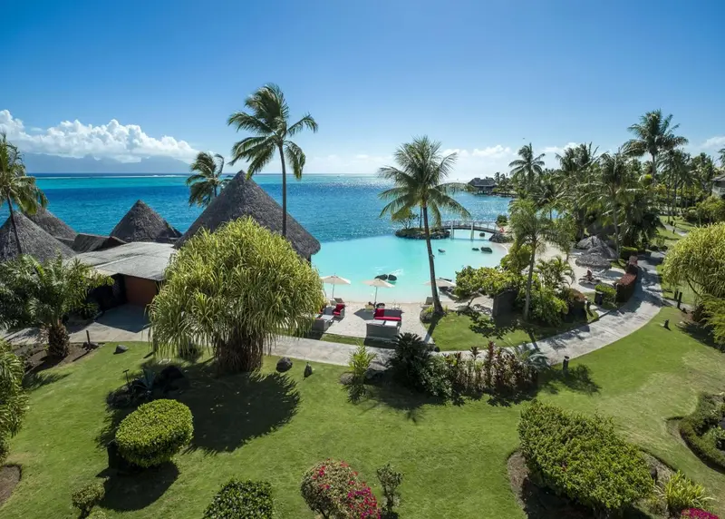 Vue aérienne d'un complexe hôtelier tropical avec des bungalows sur pilotis et une piscine turquoise.