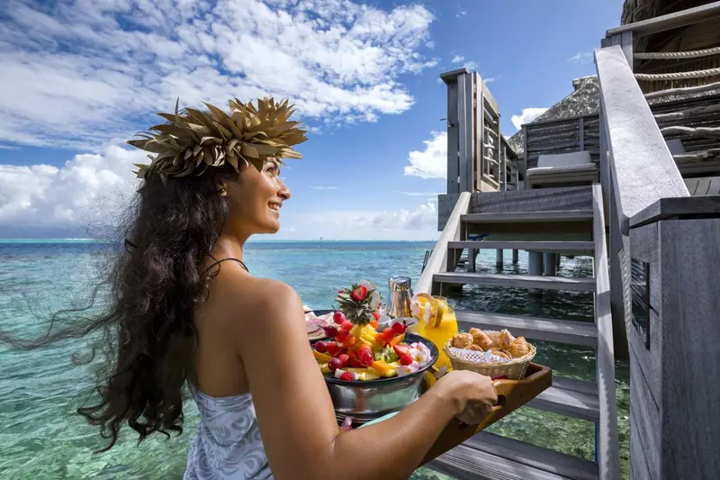Une femme souriante porte un plateau de fruits et de viennoiseries sur une terrasse au-dessus de l'eau.