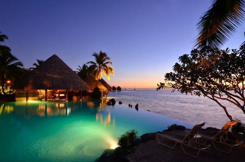 Bungalows sur pilotis illuminés au crépuscule avec piscine turquoise.