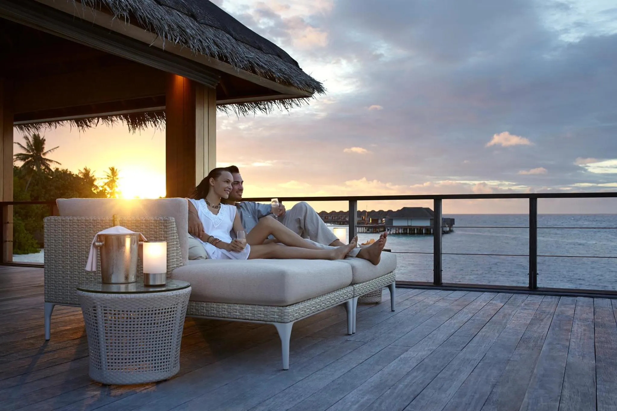 Un couple se détend sur une terrasse en bois face à la mer au coucher du soleil.