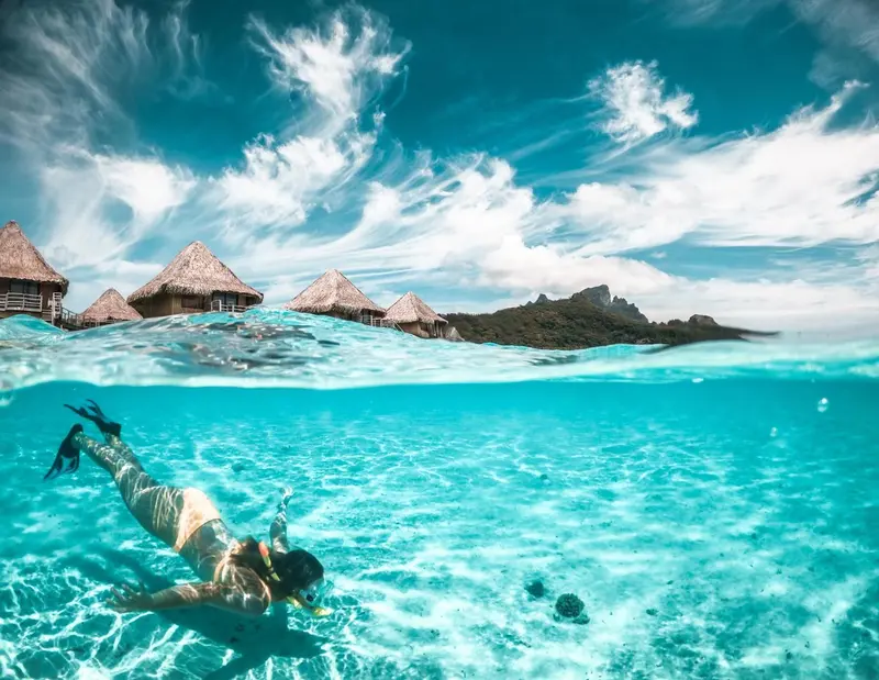 Une personne nage en surface dans une eau turquoise claire, avec des bungalows sur pilotis en arrière-plan.