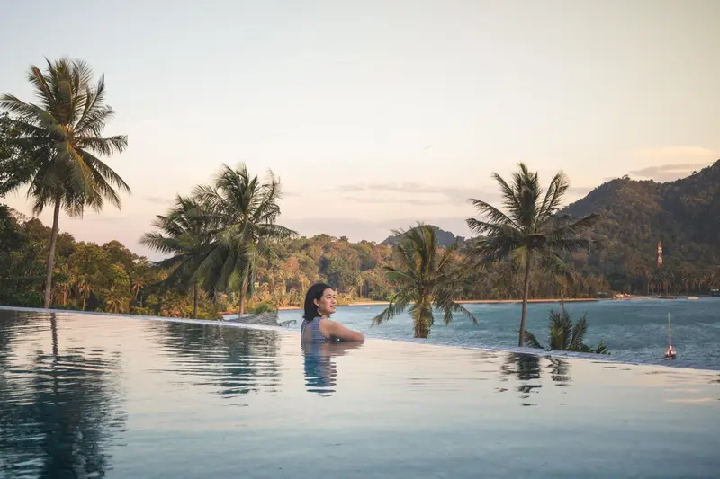 Une femme se tient dans une piscine à débordement avec une vue sur la mer et la végétation.