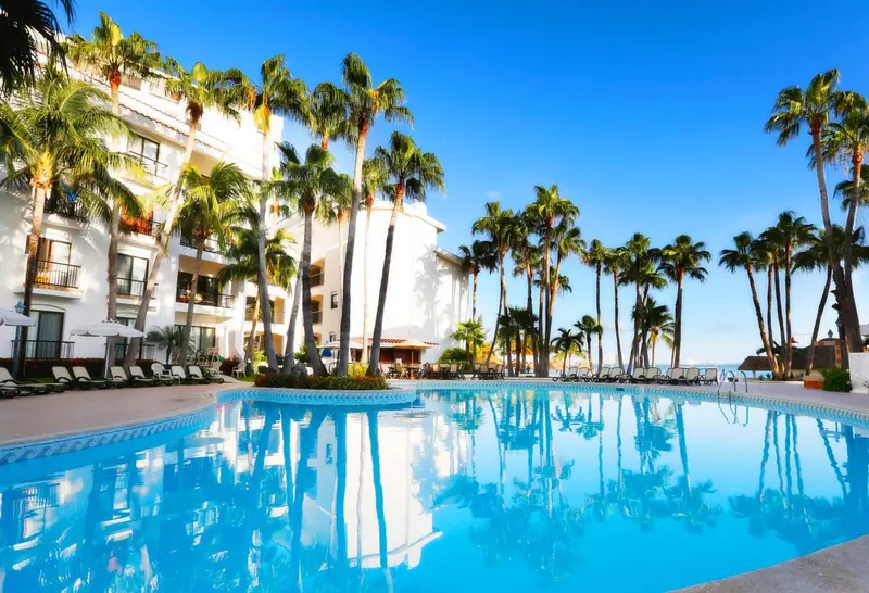 Vue d'une piscine turquoise bordée de palmiers et d'un bâtiment blanc.