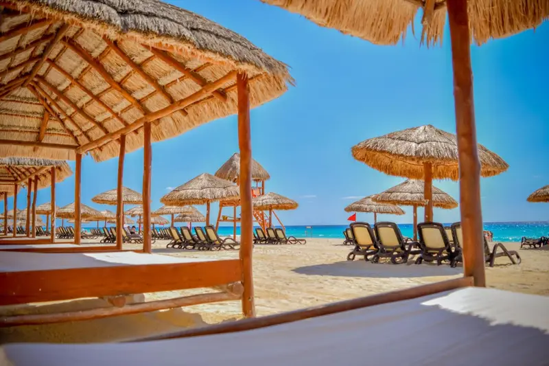 Transats et parasols de plage sous des toits de chaume, vue sur la mer.