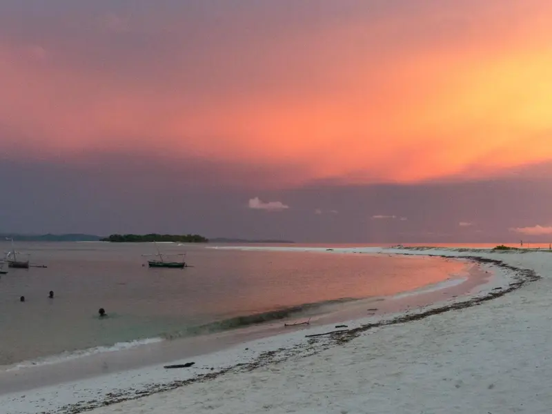 Une plage de sable blanc s'étend sous un ciel orangé au coucher du soleil.