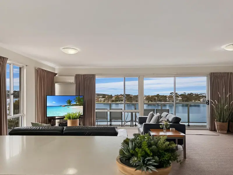 Un salon lumineux avec un grand téléviseur face à de larges baies vitrées donnant sur un paysage côtier.