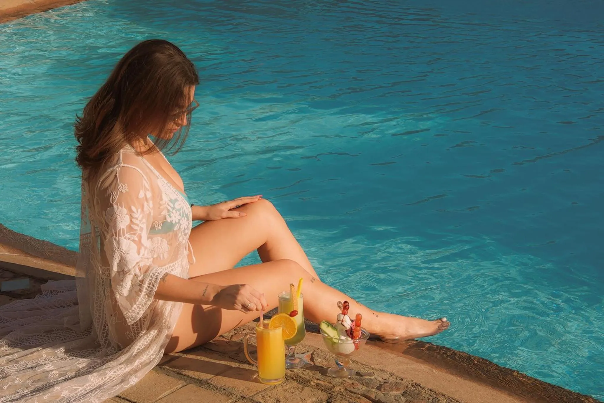 Une femme assise au bord d'une piscine avec des boissons colorées.
