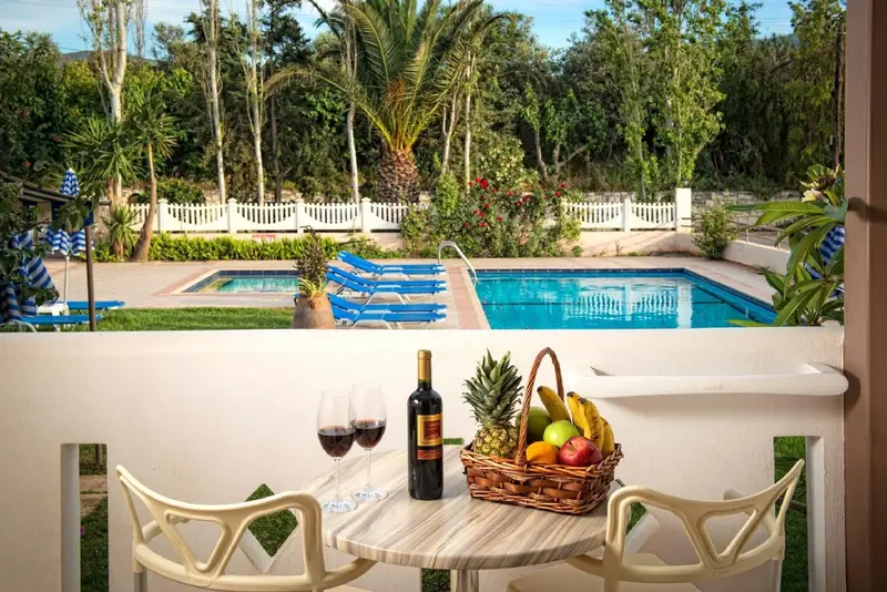 Une table avec une bouteille de vin et des fruits est mise en scène devant une piscine et un jardin luxuriant.