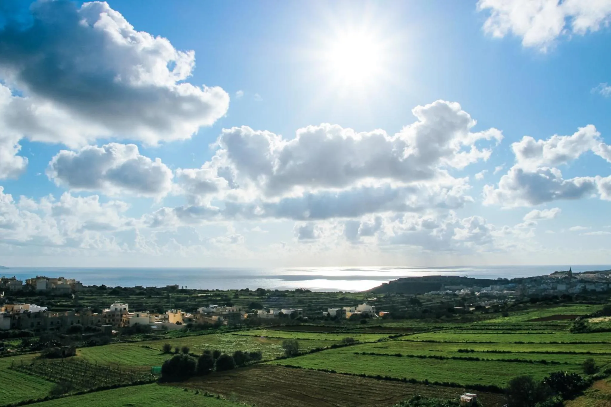 Paysage rural avec champs verdoyants et vue sur la mer sous un ciel nuageux.