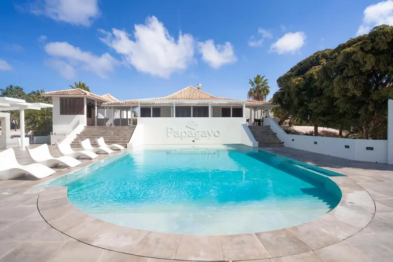 Une piscine rectangulaire bordée de chaises longues blanches devant un bâtiment blanc.