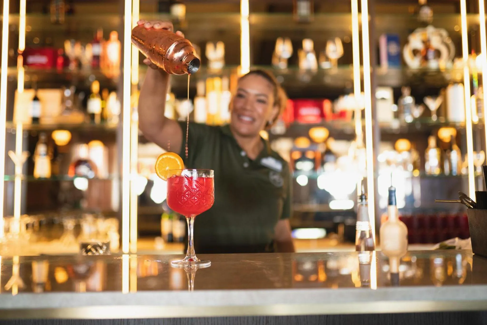 Une personne prépare un cocktail derrière un comptoir de bar illuminé.