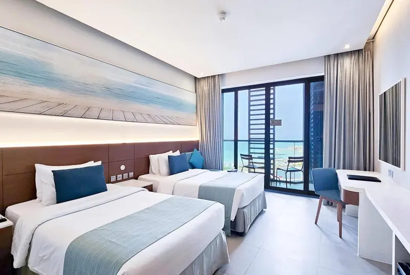 Chambre lumineuse avec deux lits et vue sur la mer depuis un balcon.