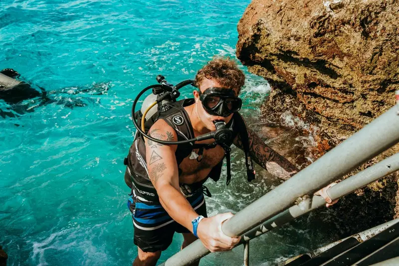 Un plongeur équipé remonte une échelle métallique au-dessus de l'eau turquoise.