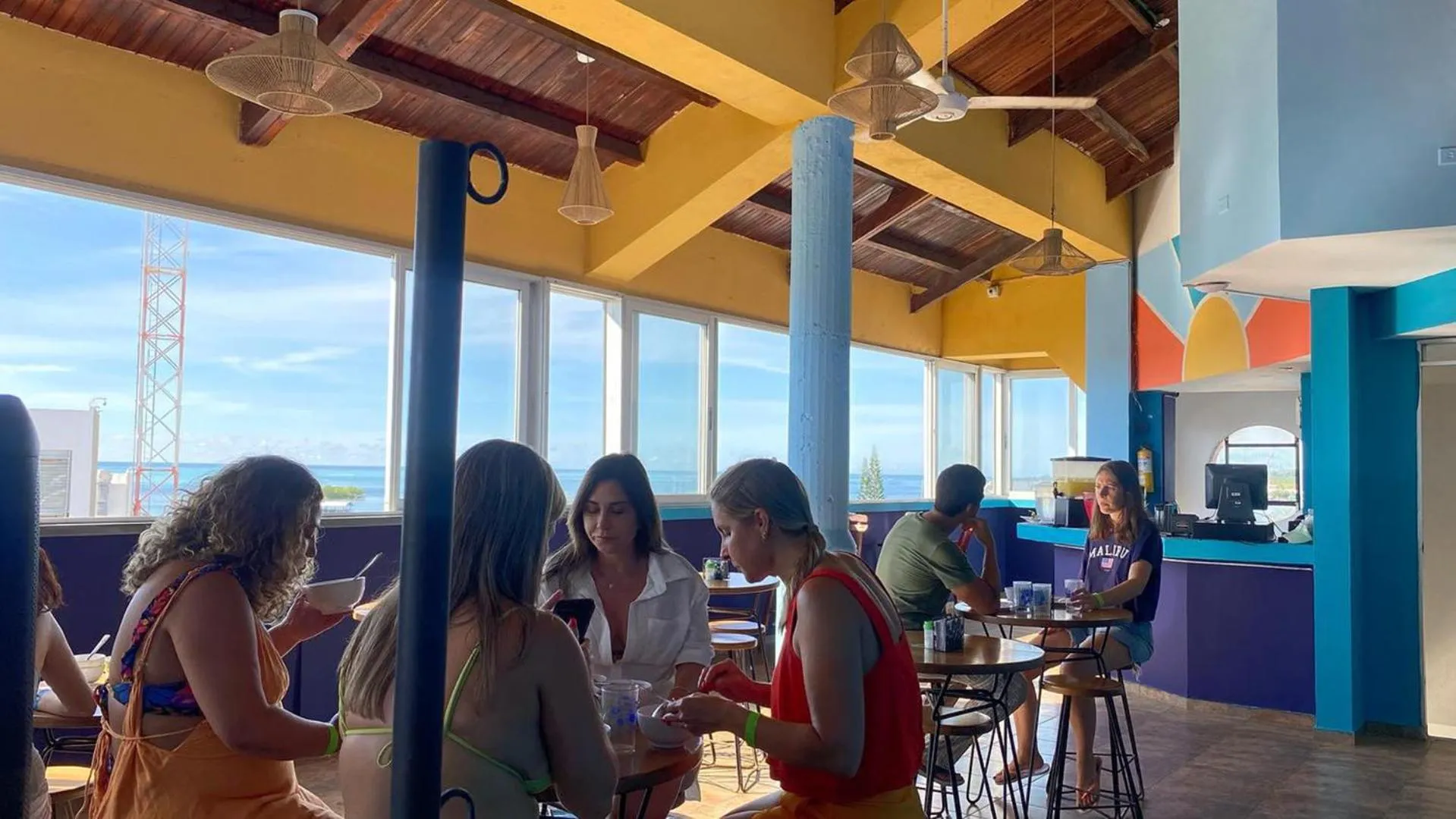 Des clients sont assis à des tables dans une salle à manger lumineuse avec vue sur la mer.