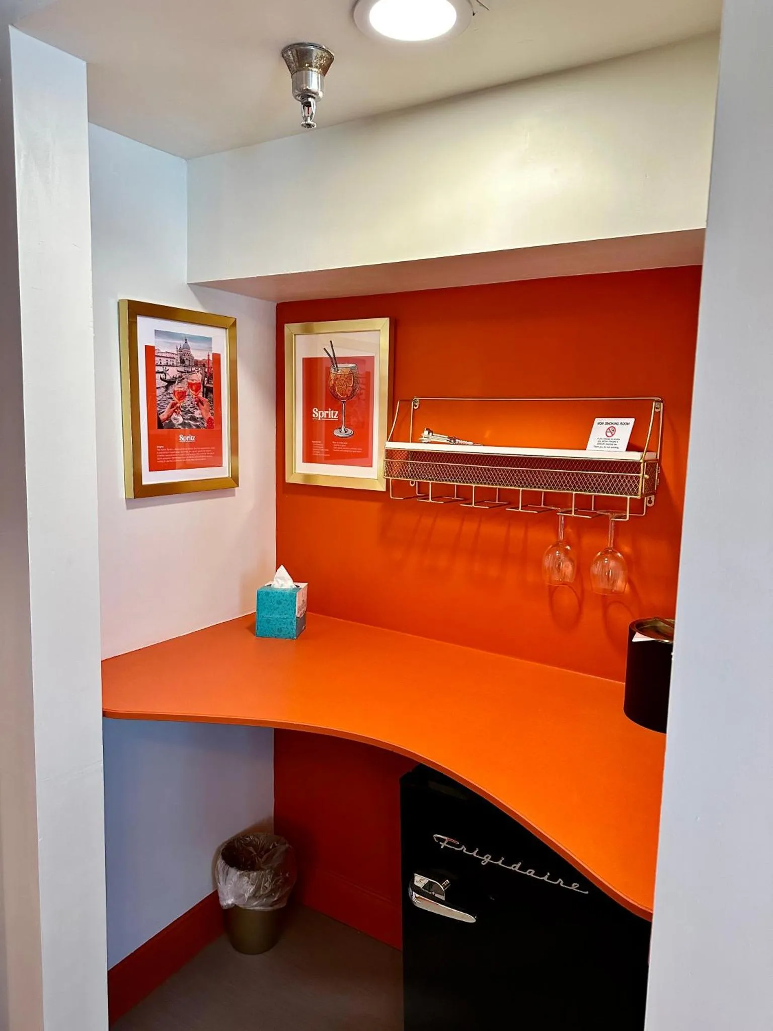 Un coin bar orange avec un réfrigérateur noir et des verres suspendus.