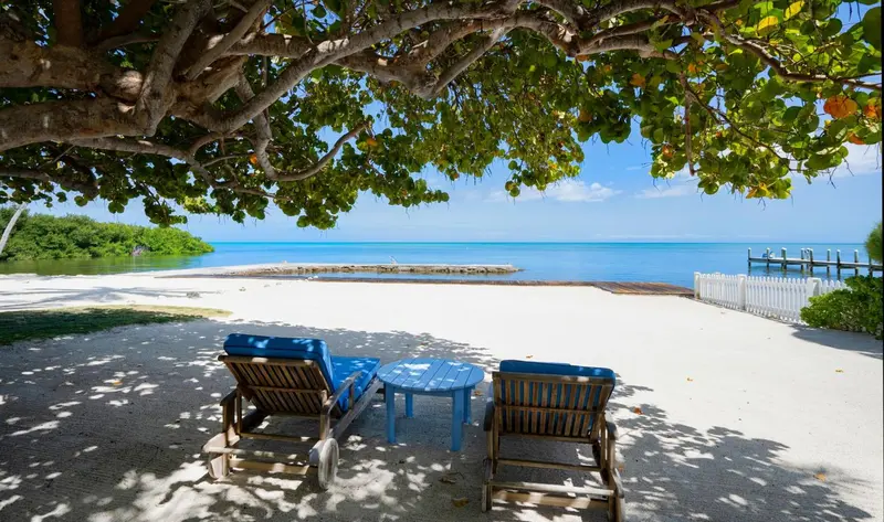 Deux transats bleus et une petite table basse sur une plage de sable blanc bordée d'arbres.