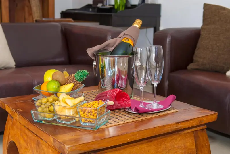 Une table basse en bois présente une composition de fruits, de snacks et une bouteille de champagne avec deux flûtes.
