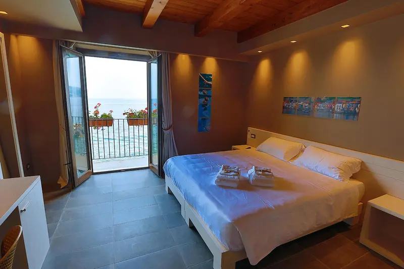 Chambre lumineuse avec un lit blanc et une vue sur la mer par de grandes portes-fenêtres.