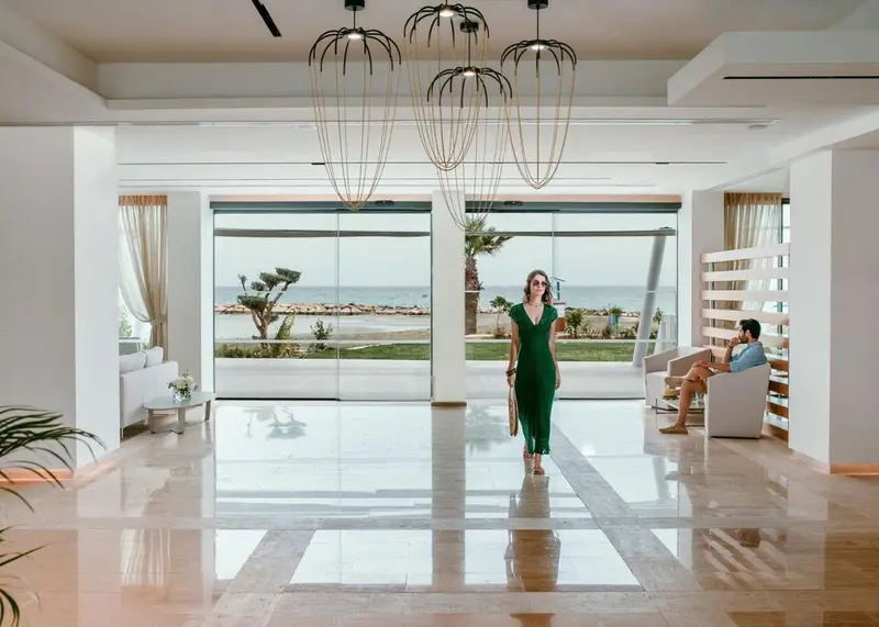 Une femme en robe verte traverse un hall d'accueil lumineux avec vue sur la mer.