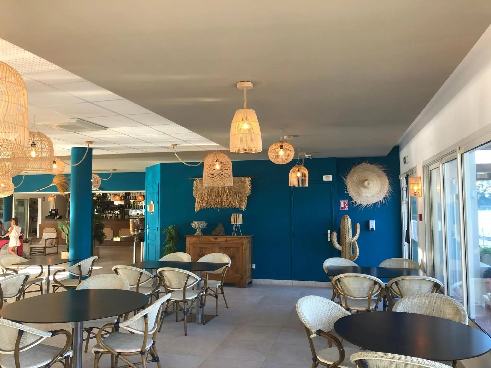 Une salle lumineuse avec des tables et des chaises est décorée de suspensions en osier et d'un mur bleu turquoise.