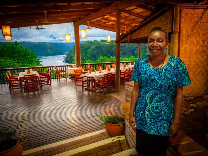 Une femme souriante se tient devant une terrasse de restaurant avec vue sur un paysage montagneux.