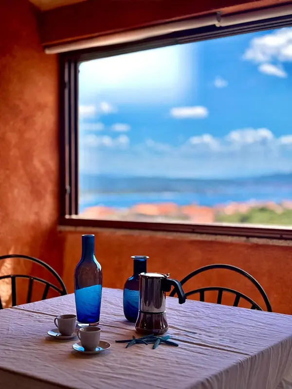 Une table avec une cafetière et des tasses est placée devant une grande fenêtre donnant sur la mer.