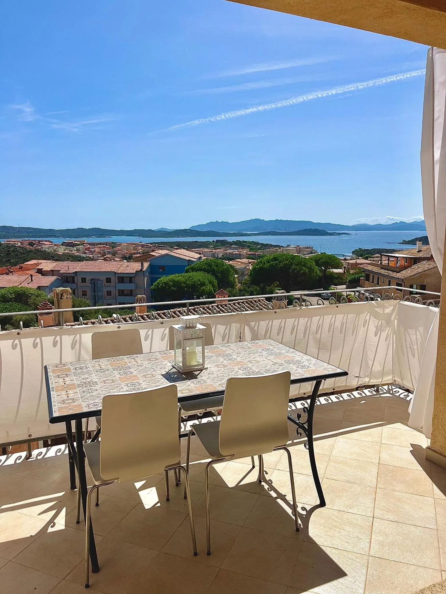 Une table et des chaises se trouvent sur une terrasse ensoleillée avec vue sur la mer et les collines.