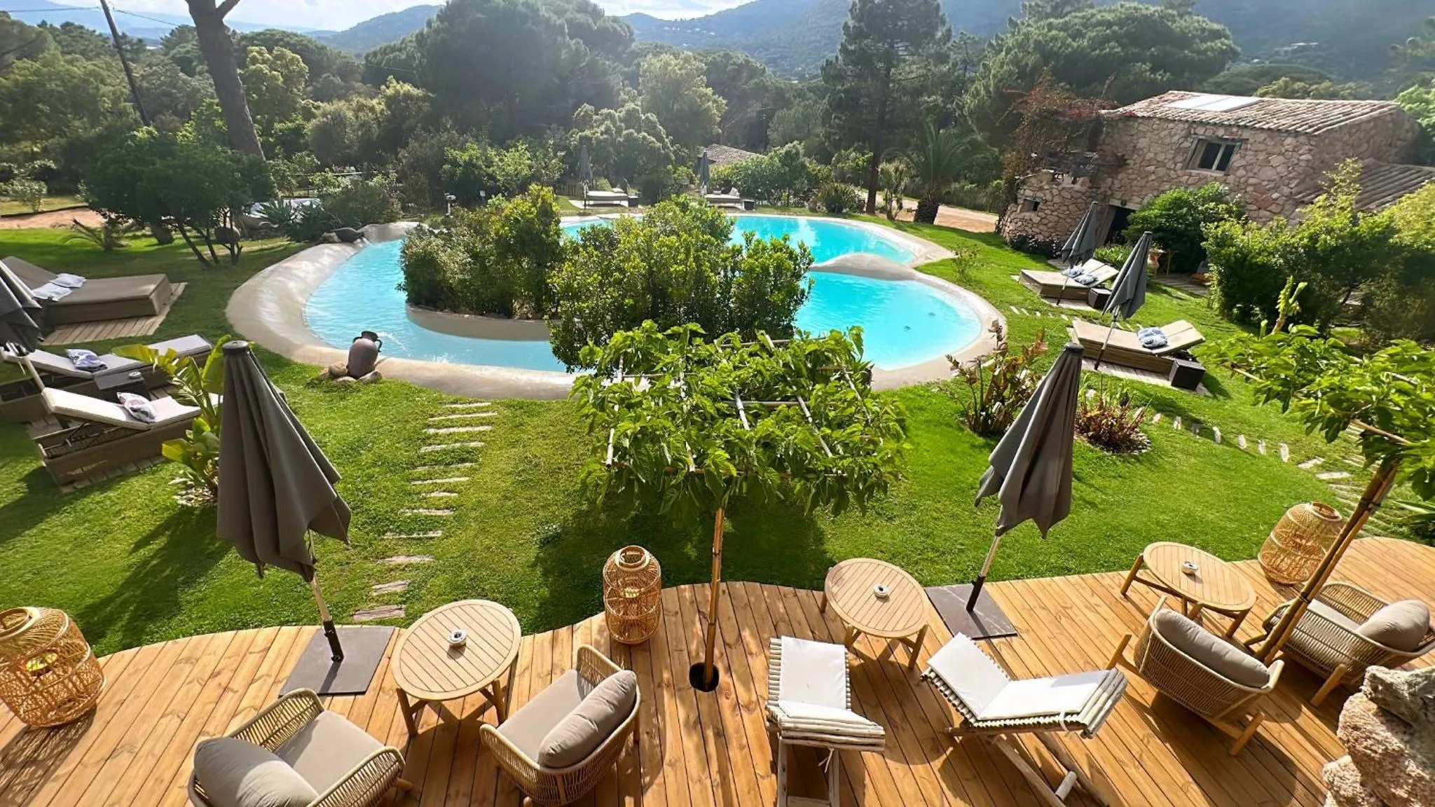 Terrasse en bois avec mobilier de détente donnant sur une piscine et un jardin verdoyant.