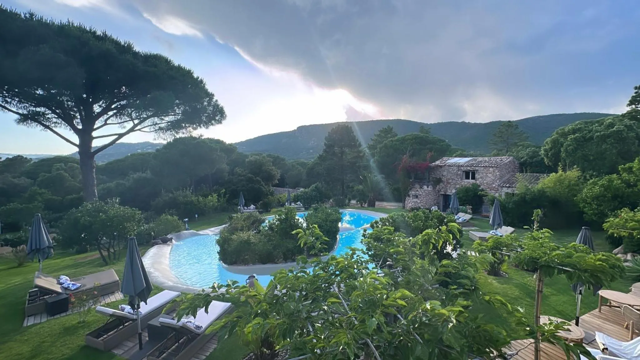 Vue d'une piscine avec des transats et des parasols dans un jardin verdoyant.