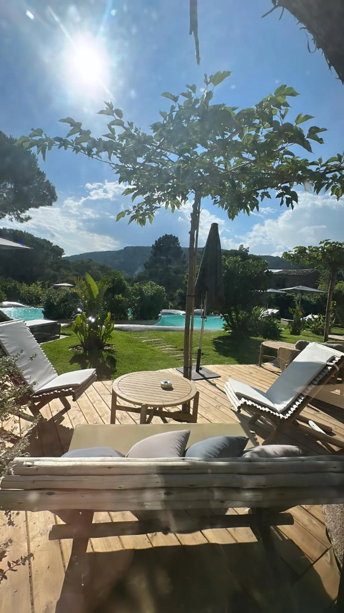 Terrasse en bois avec mobilier de détente et piscine au loin.