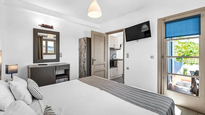 Une chambre lumineuse avec un lit fait et un espace cuisine en fond.