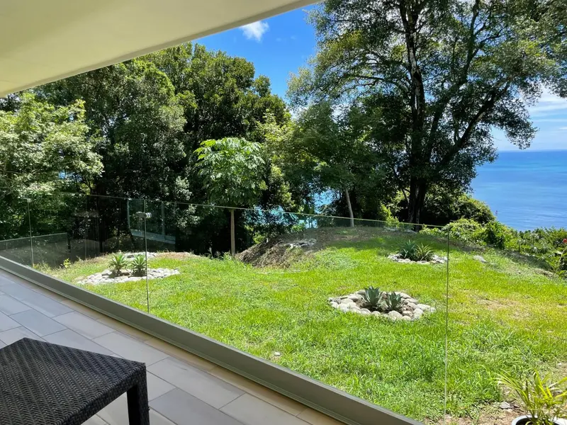 Terrasse carrelée avec garde-corps en verre donnant sur un jardin verdoyant et la mer.