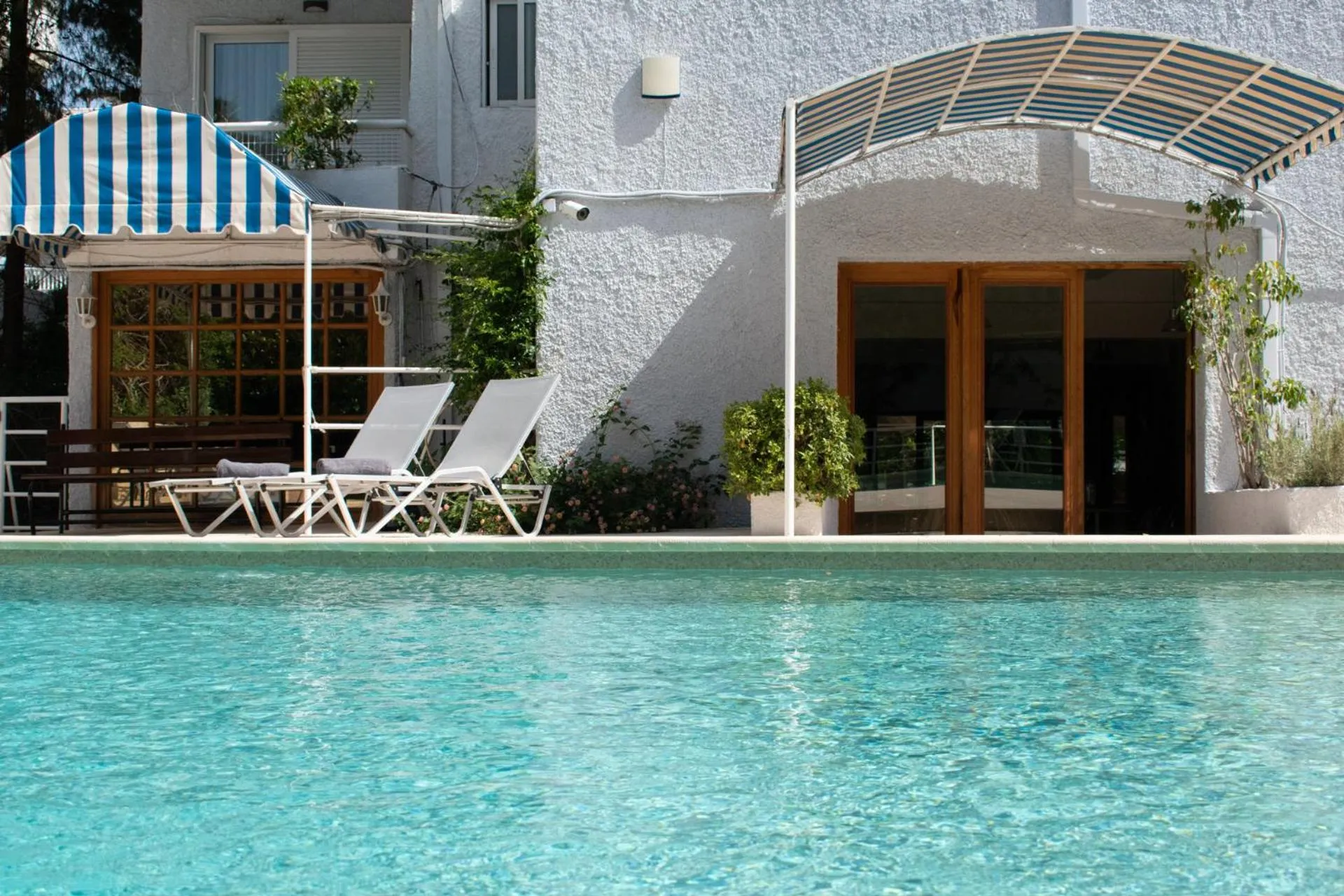 Deux transats blancs sont placés devant une piscine turquoise, sous un auvent rayé bleu et blanc.