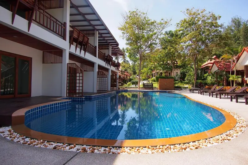 Une piscine rectangulaire bordée de galets, entourée d'une végétation luxuriante et de bâtiments blancs.