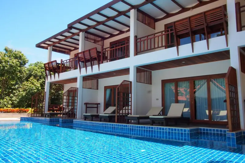 Façade d'un bâtiment blanc avec des balcons en bois donnant sur une piscine bleue.