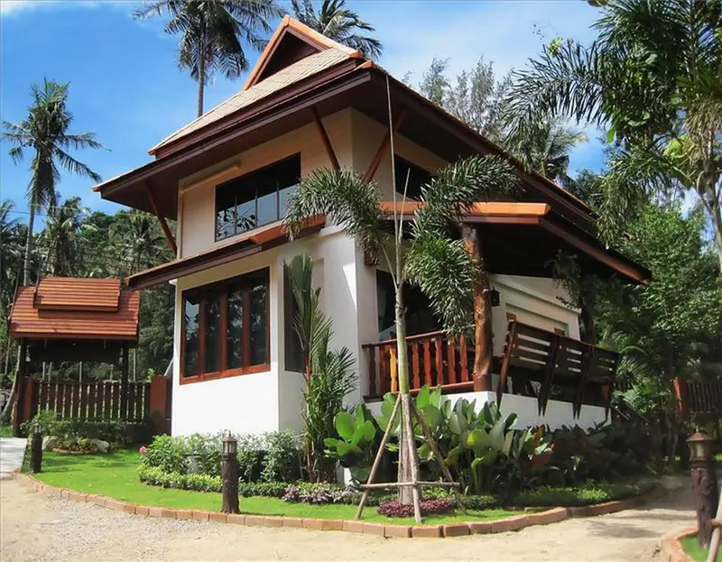 Petite maison blanche aux toits bruns, entourée de végétation tropicale.