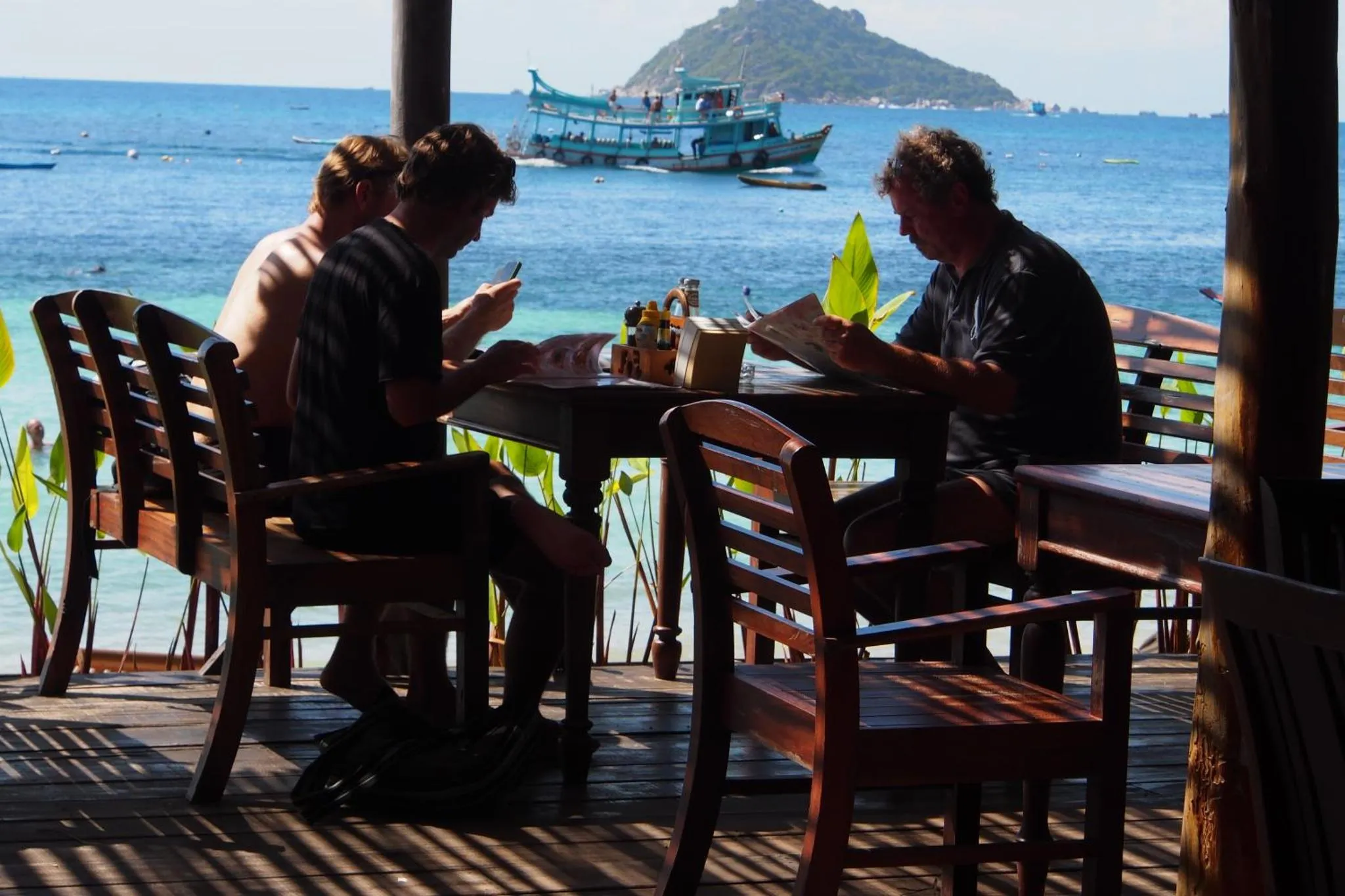 Deux personnes sont assises à une table en bois sur une terrasse ombragée face à la mer.