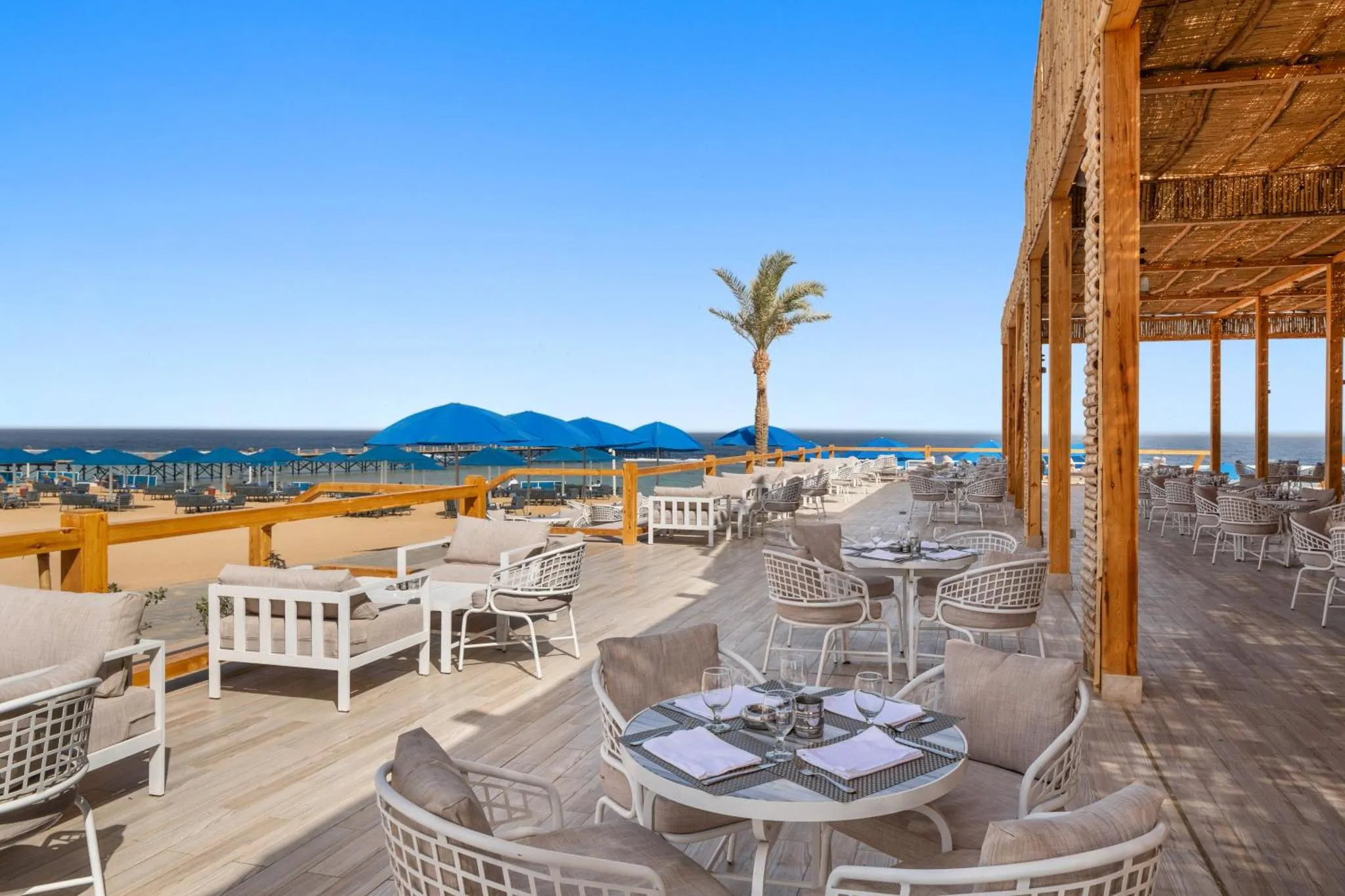 Terrasse ombragée avec tables et chaises donnant sur une plage et la mer.